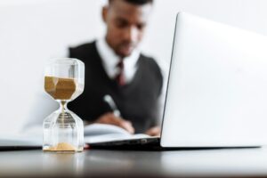 A man in black suit jacket sitting beside table with a laptop and a egg timer on the desk.