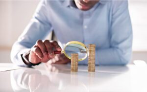 An analyst holding a magnifying glass up to two piles of coins that represent a Pay Gap. One stack is higher than the other.