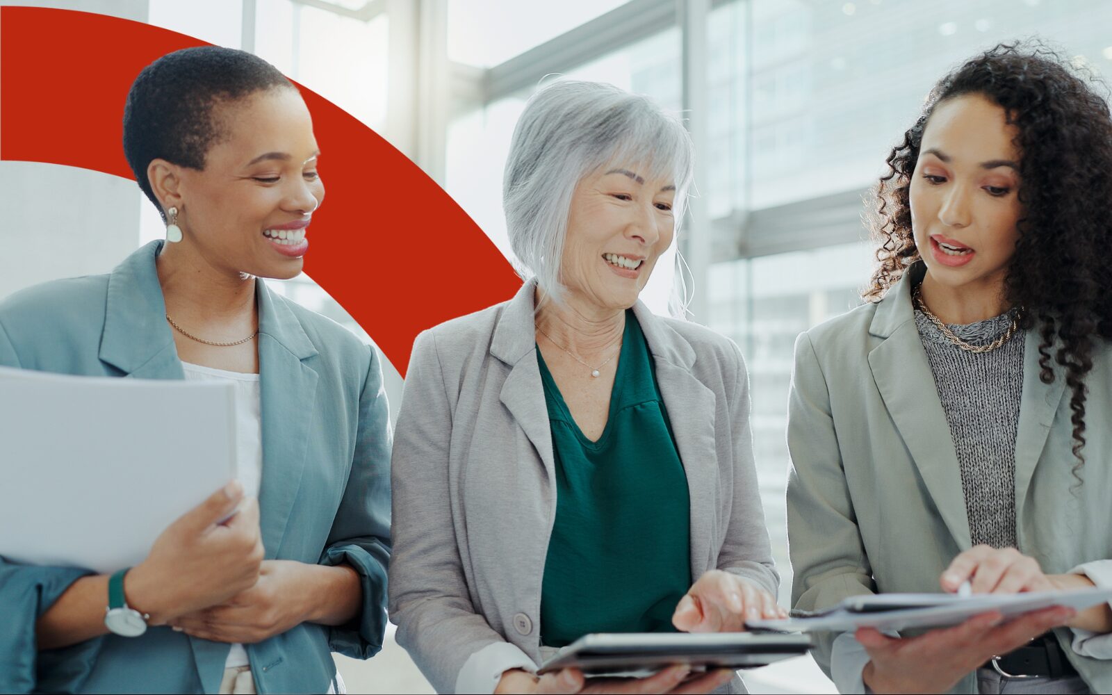 A photo of three women standing together while looking at each others notes, they are smiling and wearing grey, blue and greens.