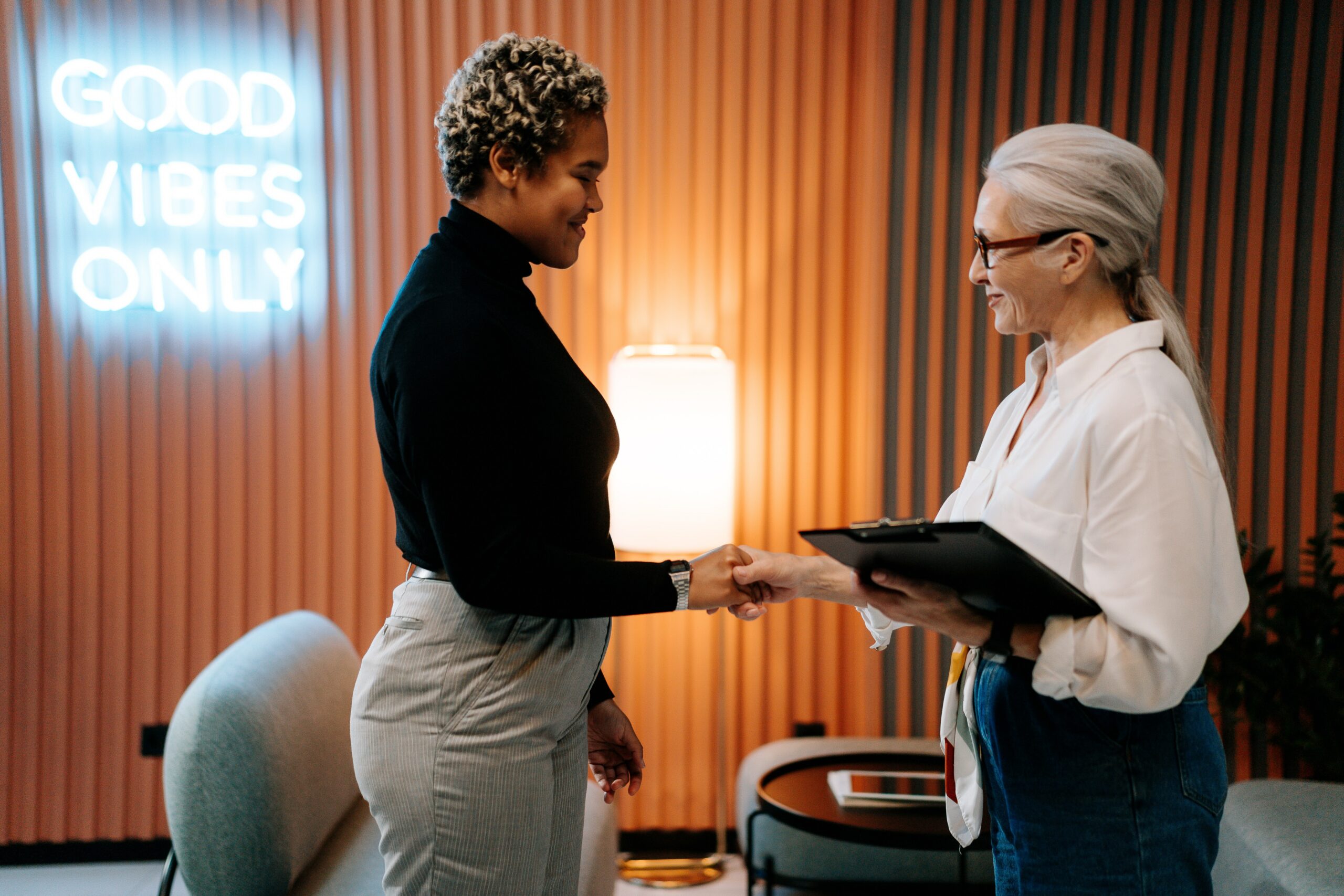An older feminine presenting White person shaking hands with a younger feminine presenting Black person in an office environment