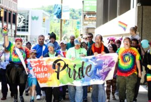 A photo of a group of people walking towards the camera with a rainbow sign that says 'Pride' in the centre in black text, and on the other side 'Ohio Pride' in white text.